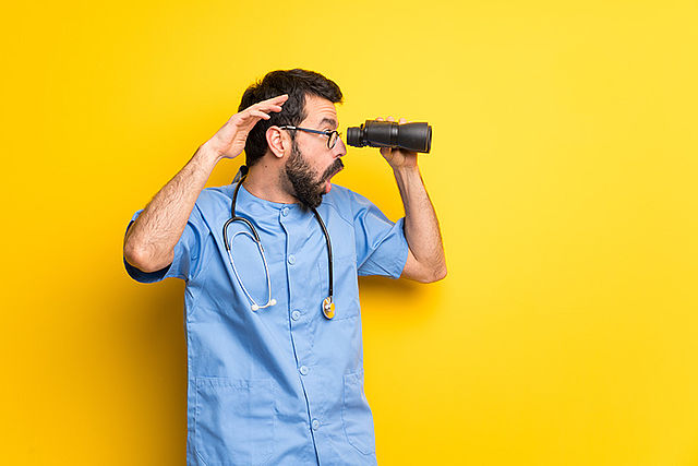 Ein Mann in blauer Pflegekleidung steht vor einer gelben Wand und schaut erstaunt durch ein Fernglas.