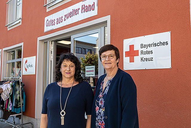 Zwei Frauen vor dem Eingang eines BRK-Kleiderladens in Eggenfelden. Im Hintergrund das Logo des Roten Kreuzes.