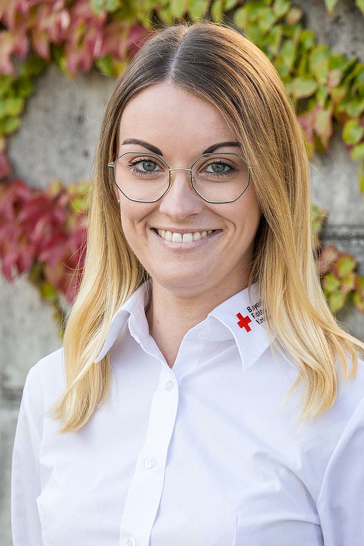 Tatjana Hammerschmidt, Assistenz der Geschäftsführung, steht vor einer Wand mit Efeu. Sie hat blondes, langes Haar und trägt eine Brille und ein weißes Hemd mit Rotkreuz-Logo am Kragen.