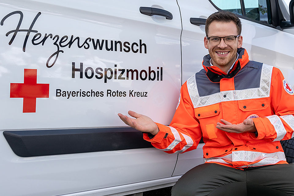 Martin Frank kniet in orangener BRK-Einsatzkleidung neben dem Herzenswunsch Hospizmobil und deutet auf dessen Logo. Er lächelt freundlich.