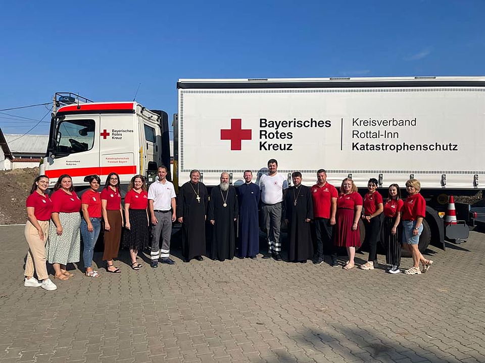 Der große Wechsellader mit dem Logo des BRK steht im Hintergrund. Im Vordergrund Mitglieder des rumänischen Roten Kreuzes und der rumänisch Orthodoxen Kirche und zwei ehrenamtliche Helfer des BRK-Kreisverbandes Rottal-Inn