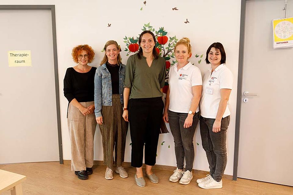 5 Mitarbeiterinnen des BRK stehen vor einer mit einem Apfelbaum bemalten Wand und schauen freundlich lächelnd in die Kamera.